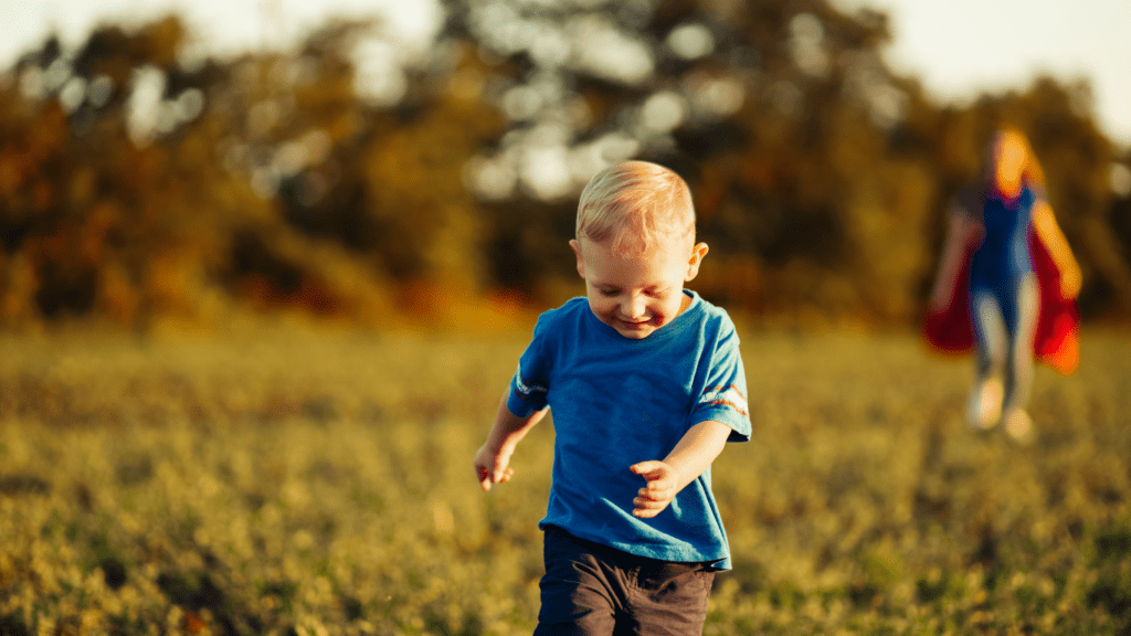 The Myth of the supermom: A mom with a cape stands behind a young toddler with blonde hair as he runs through an open field. 