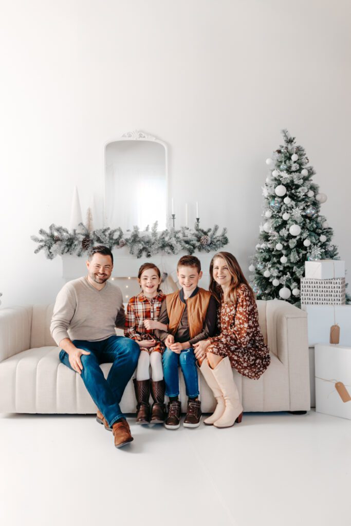 Family posing in a cozy studio setting, surrounded by holiday decorations, including twinkling lights and a beautifully decorated Christmas tree.